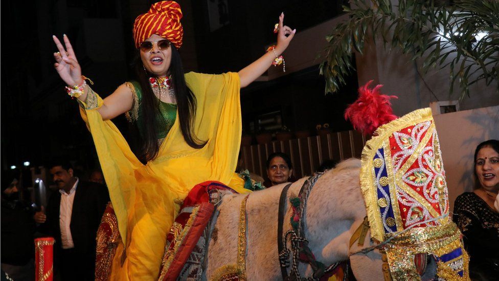 Woman riding a decked up horse, dressed in yellow sari, wearing a pagdi, with both her hands up in the air. One women looking up at her. Another woman laughing. Other people faintly visible. 
