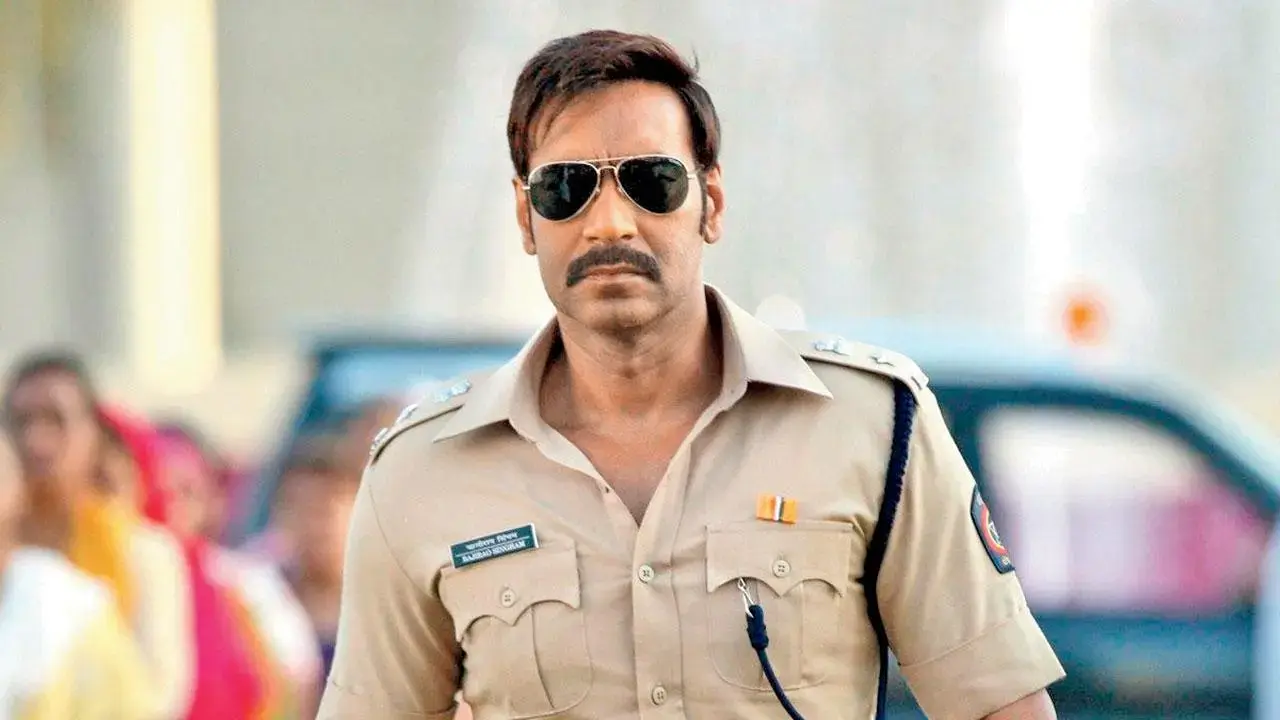 Ajay Devgn looking stern. Wearing aviators and a khaki police uniform. The background is in soft focus showing some people on the left and a car behind him.