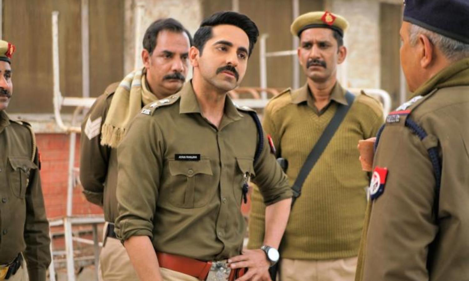 Ayushmann Khurrana is in the centre in a khakhi police uniform, with his sleeves rolled up and left hand on hip. He is talking to another police officer whose back is facing the camera. There are three other policemen in the background standing and listening to them.