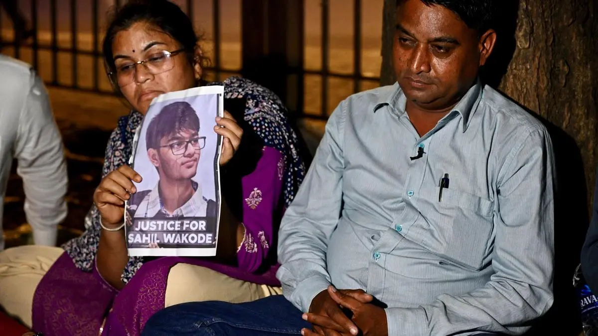 A woman wearing glasses and a purple and cream salwar kameez and dark printed dupatta holds up a poster with the photo of a young man wearing glasses, a light colour shirt with collars and what looks like a dark sweater. She is seated next to a man in light blue shhirt.