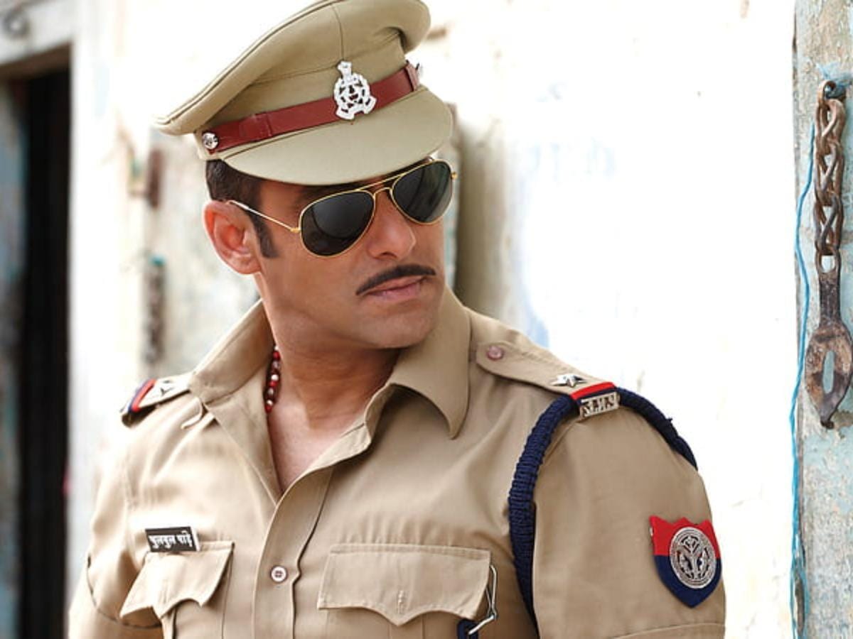 Salman Khan looking to his left; wearing a khakhi police uniform and aviators. Looking stern. Standing against a wall with an open door and something metallic hanging on the right side.