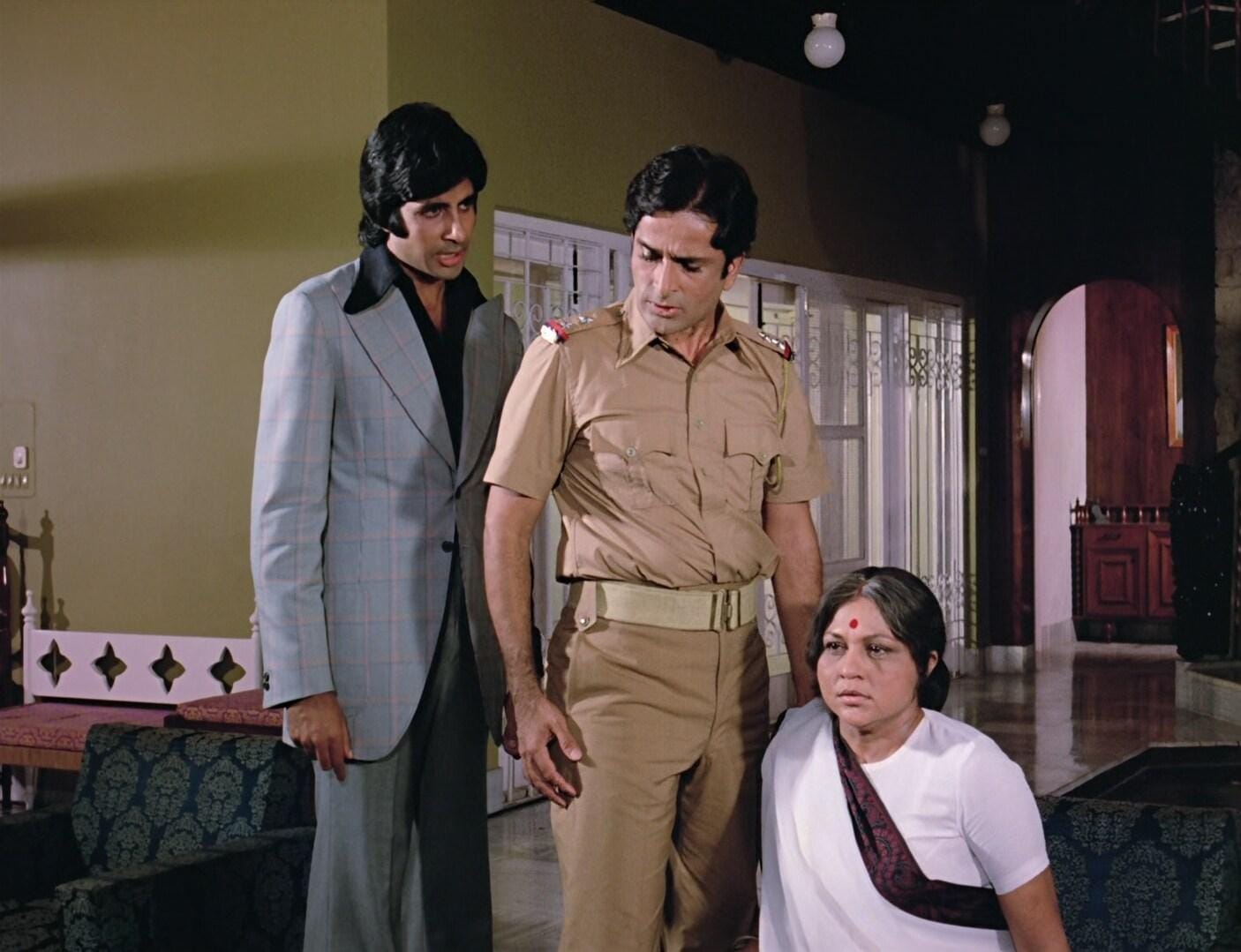 Scene from the film Deewar. In an indoor space that looks like a living room, Amitabh Bachchan is standing in a grey business suit talking to Shashi Kapoor standing in the middle wearing a khakhi police uniform. Seated to Shashi Kapoor's right is Nirupa Roy wearing a bindi and a white saree with a dark border.