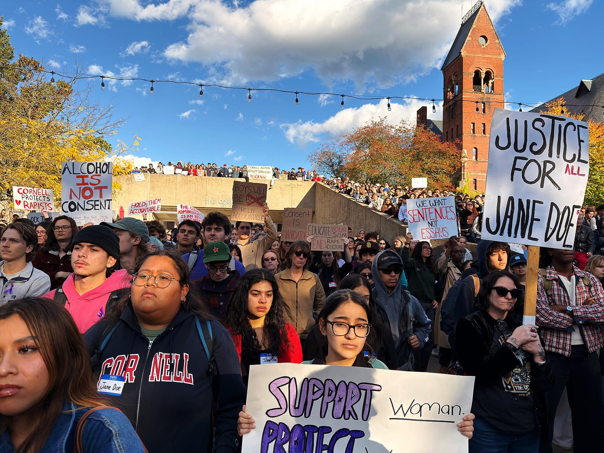 Image of the front part of a large crowd of protesters, some holding placards reading "Alchohol is not consent" and "Justice for (All) Jane Doe(s)." In the background the sky is blue with patches of white clouds. Cornell's tower can be seen on the right side.