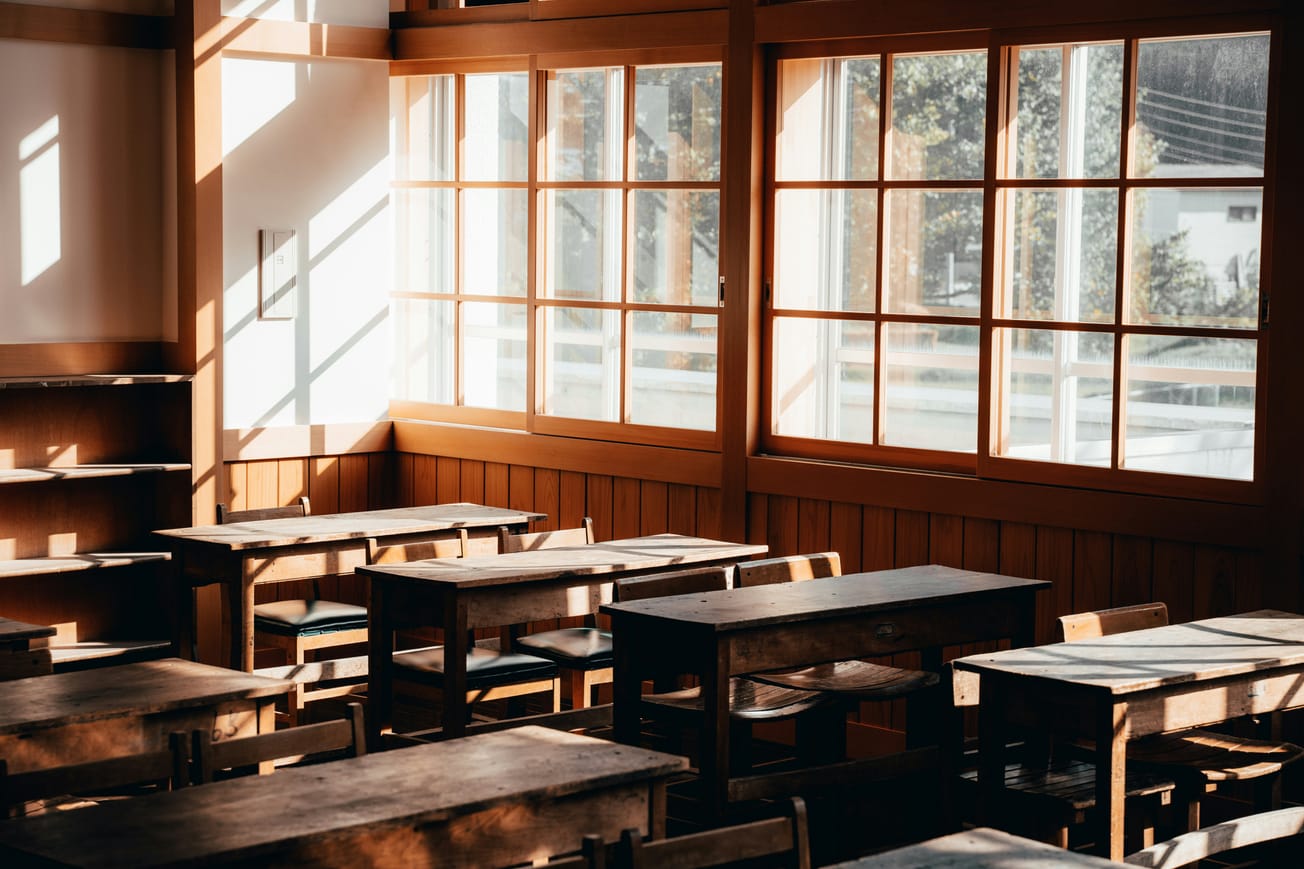 An empty classroom with desks and chairs for two students to sit together. In the background, large windows with sun filtering in to the room an on to the desks and chairs.