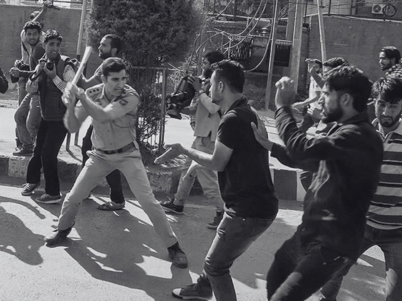 A police officer in uniform swings a baton at a man wearing a dark T-shirt and trousers. Another man, also in dark clothes. Both men in defensive stance. Several other men in the background.